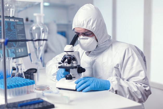 Microbiologist with protection glasses using microscope dressed ppe suit